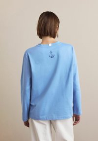 Svetlo modra majica z dolgimi rokavi iz bombaža, z majhno mornarsko sidroveve vezenine na hrbtni strani, z okroglim izrezom in sproščenim krojem.