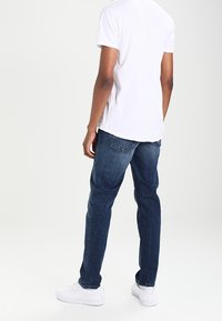 Bílé tričko s krátkými rukávy a švem na zádech, ve spojení s tmavě modrými džíny a bílými teniskami. Materiál působí hladce a volně.