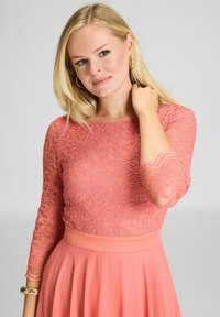 Korallinvärinen pitsipaita kolmen neljänneksen hihoilla ja aaltomaisella helmaa, paritettu yhtenäisen korallinvärisen hameen kanssa, joka on sileä ja virtaava.