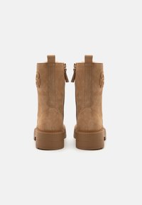 Kamakad tan suede pahkliku boots, millel on jämedad tallad, tagumine tõmblukk ja külgedel õmmeldud logo detail. Pehme vooder on nähtav ülaosas.