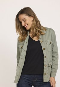 Veste bouton-unique vert clair en coton, avec deux poches poitrine, des boutons en écaille de tortue et une coupe décontractée sur un haut en V noir.