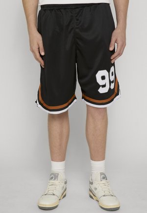 Crne košarkaške kratke hlače s narančastim i bijelim prugama, broj 99 na lijevoj nozi, nošene s bijelim čarapama i bijelim tenisicama, stojeći na jednobojnoj pozadini.