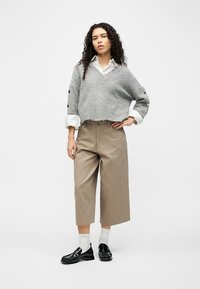 Γυναίκα με σγουρά μαλλιά που φοράει γκρι cropped πουλόβερ πάνω από λευκό πουκάμισο, μπεζ φαρδιά cropped παντελόνια, λευκές κάλτσες και μαύρα loafers.