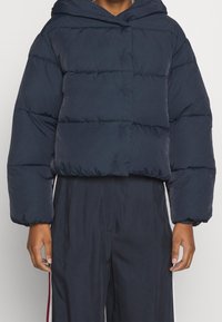Doudoune courte bleu marine avec capuche, présentant un design matelassé, des poignets élastiques et une texture lisse. Adaptée aux temps froids.