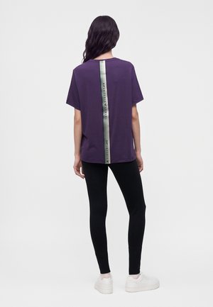 Femme debout, de dos, portant un t-shirt violet avec une bande verticale argentée et un legging noir, sur un fond clair uni.