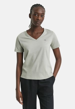 T-shirt di cotone verde chiaro con scollatura a V e maniche corte. Presenta un piccolo logo ricamato sul petto. Indossato con pantaloni neri.