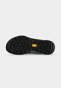 Semelle de chaussure noire et robuste avec des rainures profondes et un petit logo jaune au centre, présentée sur un fond clair uni.