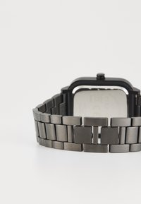 Montre rectangulaire noire avec un bracelet en maillons métalliques, présentant un mélange de surfaces mates et brillantes, et un boîtier carré noir.