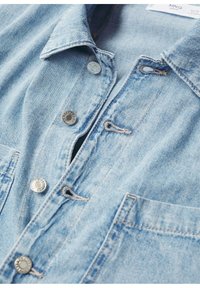 Veste en denim bleu clair avec boutons-pression métalliques, boutonnières, col et poche avant, montrant une partie de l'étiquette du vêtement.