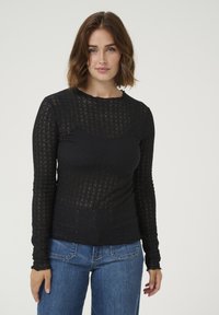 T-shirt noir à manches longues avec un motif texturé, col rond et coupe ajustée. Associé à un jean bleu, mettant en valeur un look décontracté.