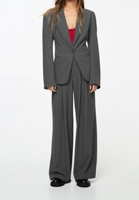 Costume gris sur mesure avec pantalon à jambes larges et blazer ajusté. Le tissu semble lisse, avec une fermeture à un bouton sur le devant.