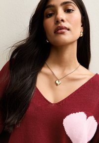 Maroon sweater with a pink heart pattern on the chest, paired with a gold heart pendant necklace on a silver chain. Smooth texture.