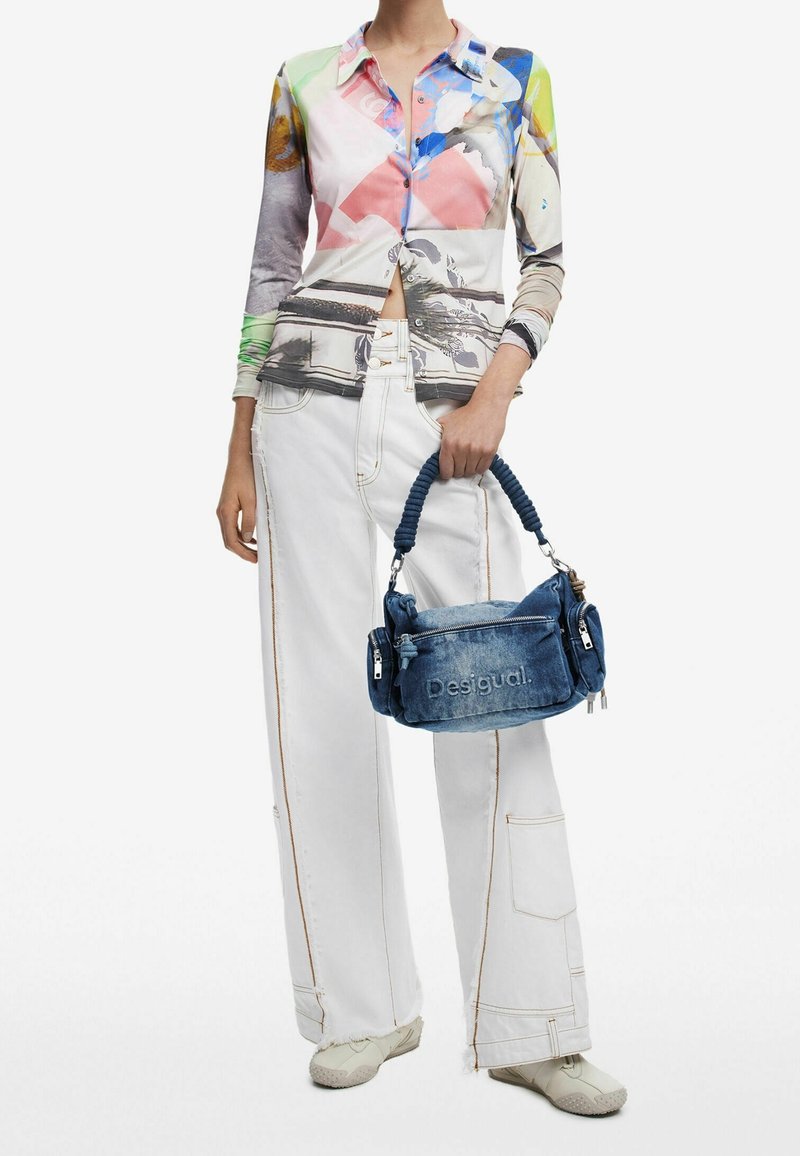 Chemise à manches longues colorée avec des motifs abstraits, associée à un pantalon large blanc. Tient un sac en denim bleu avec une anse tressée et un logo.