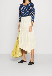 Marineblaues Oberteil mit bunten Polka-Dots, creamfarbene plissierte Rock, schwarze Mules und eine strukturierte gelbe Tasche. Lässige Kleidung mit Schwerpunkt auf Schichten.