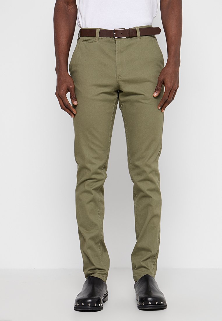 Olivgröna byxor i bomullsblandning, slim fit-design, med två framfickor, brunt läderbälte och svarta ankelboots.