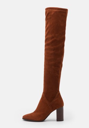 Botte haute en daim marron avec un bout pointu et un talon bloc superposé, s'étendant au-dessus du genou.
