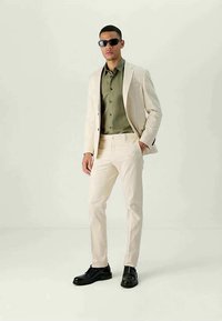 Man wearing cream suit, olive green shirt, black shoes, and black sunglasses standing against plain light background.