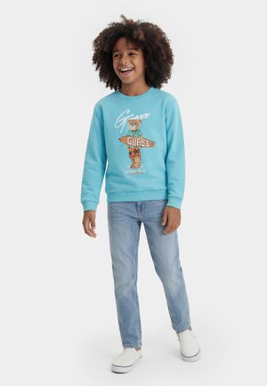 Bambino sorridente con capelli ricci indossa una felpa blu con un orso che tiene una tavola da surf, jeans azzurri e scarpe bianche slip-on.