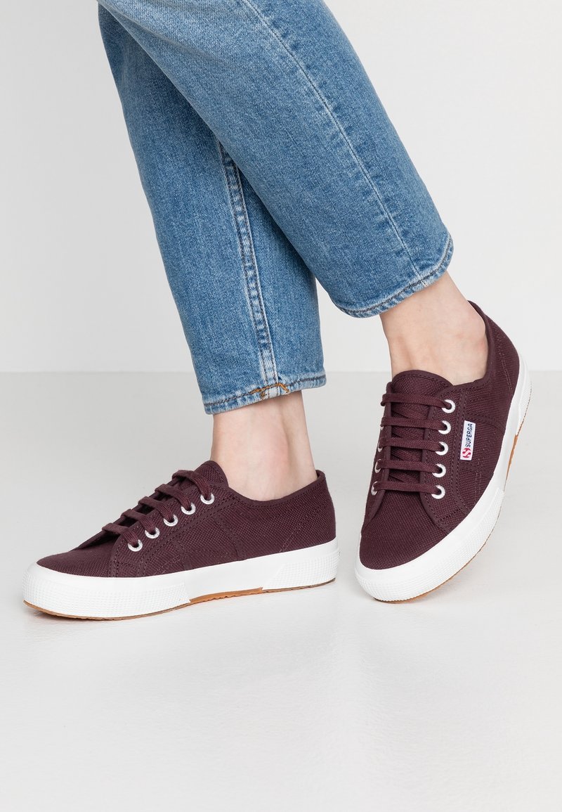 Personne portant un jean bleu et des baskets en toile violet foncé avec des semelles en caoutchouc blanches, debout sur un sol clair.