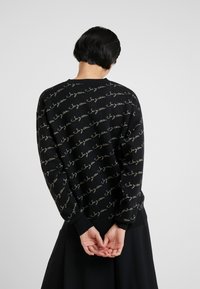 Osoba z krótkimi czarnymi włosami stoi z rękami złożonymi za plecami, ubrana w czarny sweter z powtarzającym się tekstem i czarną spódnicę.