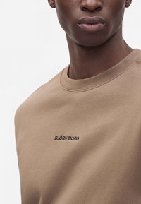 Gros plan d'une personne portant un sweat-shirt beige avec le texte "BJÖRN BORG" brodé en noir sur la poitrine.