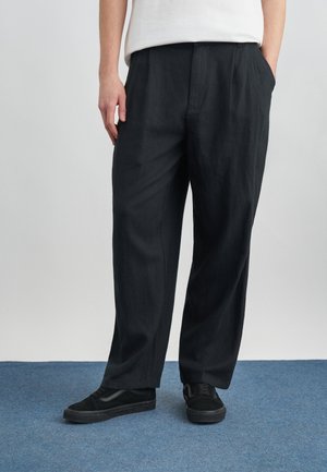 Personne portant un pantalon noir ample et des baskets noires, debout sur un tapis bleu les mains dans les poches, devant un fond clair uni.