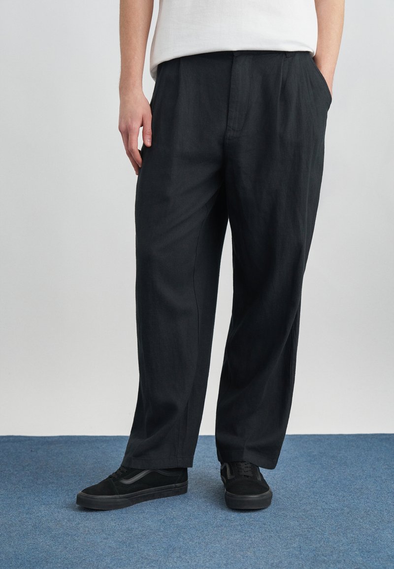 Άτομο που φοράει φαρδιά μαύρη παντελόνια και μαύρα αθλητικά παπούτσια, στέκεται σε μπλε χαλί με τα χέρια στις τσέπες, μπροστά σε απλό φωτεινό φόντο.