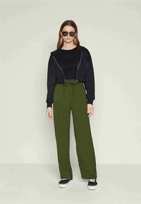 Γυναίκα με μαύρο κοντό φούτερ, μαύρη μπλούζα, ελαιόλευκα παντελόνια φαρδιάς γραμμής, μαύρα γυαλιά ηλίου και μαύρα αθλητικά παπούτσια με λευκές σόλες.