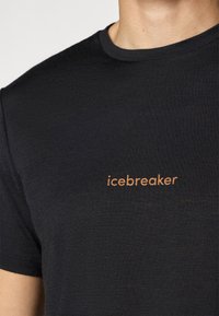 Close-up van een persoon die een zwart T-shirt draagt met het "icebreaker"-logo in lichtoranje op de borst.