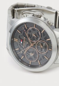 Silverfärgad klocka i rostfritt stål med en grå skeleton urtavla. Innehåller tre sub-dials, roséguld accenter och numeriska timmarkörer. Logotyp synlig.