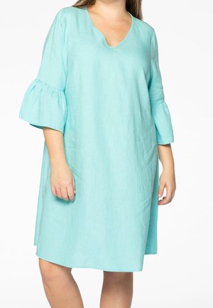 Vrouw draagt een loszittende, lichtblauwe jurk tot op de knieën met een V-hals en ruches aan de mouwen, staand tegen een effen witte achtergrond.