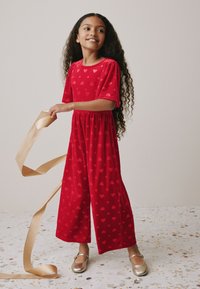 Tuta rossa con motivo a cuori, maniche corte a sbuffo, vita elastica e gambe larghe. Scarpe da balletto dorate completano l'outfit.