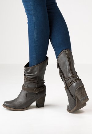 Bottes en cuir gris foncé mi-mollet avec fermetures éclair, boucles, chaînes et talons carrés portées avec un jean skinny bleu.