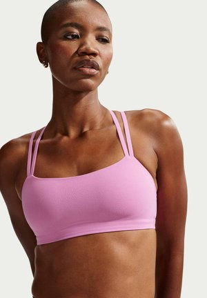 Kobieta z krótkimi włosami, ubrana w jasnofioletowy sportowy biustonosz z podwójnymi ramiączkami, patrząca w dół na jednolitym tle.