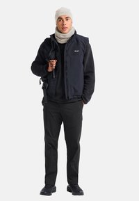 Gilet bleu foncé sur une polaire noire, bonnet en tricot beige et col en polaire, pantalon noir et chaussures robustes. Le gilet a des poches zippées.