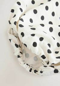 Balts audums ar melniem punktiņiem, sasiets ar elastīgām malām, veidojot apaļu, savērptu formu uz neitrālas fona.