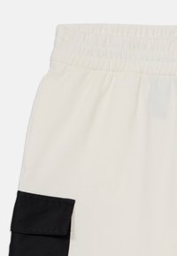 Pantaloni color crema con vita elasticizzata e tasca laterale in tessuto nero. Texture liscia con colori a contrasto e design semplice.