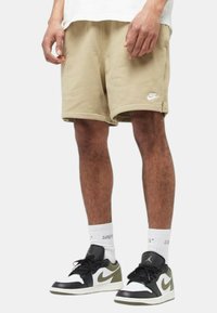 Pantaloni sportivi beige con cintura elasticizzata, tasche laterali e logo Nike bianco. Indossati con scarpe da ginnastica nere e verdi e calzini bianchi di media lunghezza.