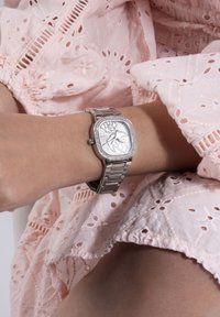 Reloj de pulsera de metal plateado con esfera cuadrada, adornado con un patrón, rodeado de pequeños cristales, combinado con una prenda texturizada de color rosa.