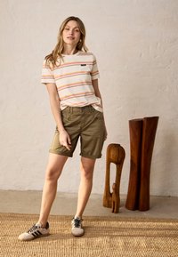 Jeune femme portant un T-shirt rayé, un short vert olive et des baskets beiges, debout à l'intérieur près de deux grandes sculptures en bois.