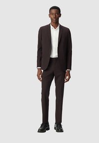 Traje marrón oscuro de corte entallado, chaqueta de un solo botonadura, pantalones a juego y zapatos negros. Camisa blanca visible bajo la chaqueta.