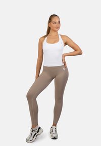 Bela halter majica in bež legice iz raztegljive tkanine, ki se ponašajo z gladko teksturo in logotipom na nogi. Urejeno s športnimi čevlji.