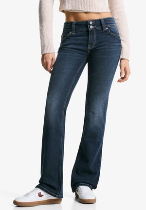 Blauwe flared jeans met een hoge taille, zilveren hardware en contrasterende stiksels. Gecombineerd met witte sneakers en een lichtroze sweater.