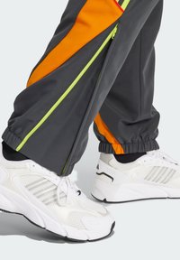 Grå træningsbukser med orange og lime detaljer, der har lynlåse og samlede manchetter. Hvide sneakers med netmateriale og støttende såler.