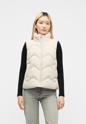 Femme aux longs cheveux blonds portant une doudoune beige matelassée sans manches sur un haut noir à manches longues et un jean gris clair, debout sur fond blanc.