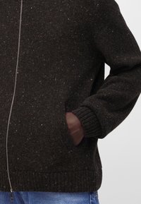 Gros plan d'une personne portant un pull en laine zippé foncé tacheté, avec une main dans la poche avant et un jean bleu visible.