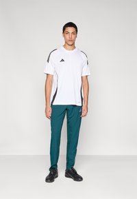 White short-sleeved athletic shirt with black accents, paired with teal tapered pants featuring side stripes and black athletic shoes.