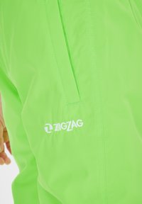 Pantaloni verde neon realizzati in tessuto leggero con una texture liscia, dotati di una tasca con zip e dettaglio del logo bianco "ZIGZAG".
