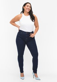 Top bianco senza maniche con scollatura alta, abbinato a jeans blu scuro a vita alta. La modella indossa scarpe con punta azzurra chiara. Sfondo neutro.