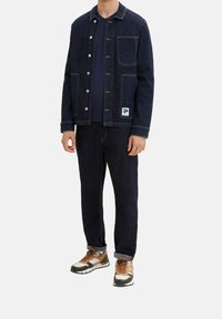 Dunkle Jeansjacke mit Knopfverschluss, vier Taschen, kontrastierenden Nähten, kombiniert mit dunkler Jeanshose und mehrfarbigen Turnschuhen.